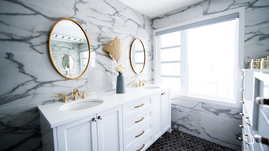 Marble-clad bathroom with a large round mirror and a smaller oval one in brass, above a double vanity