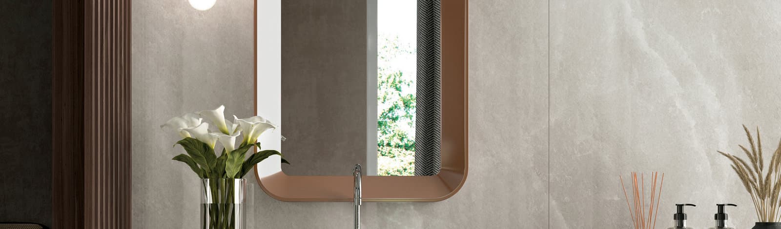 A warm bronze-framed mirror above a stone basin in a travertine bathroom, lit low with daylight through the window behind