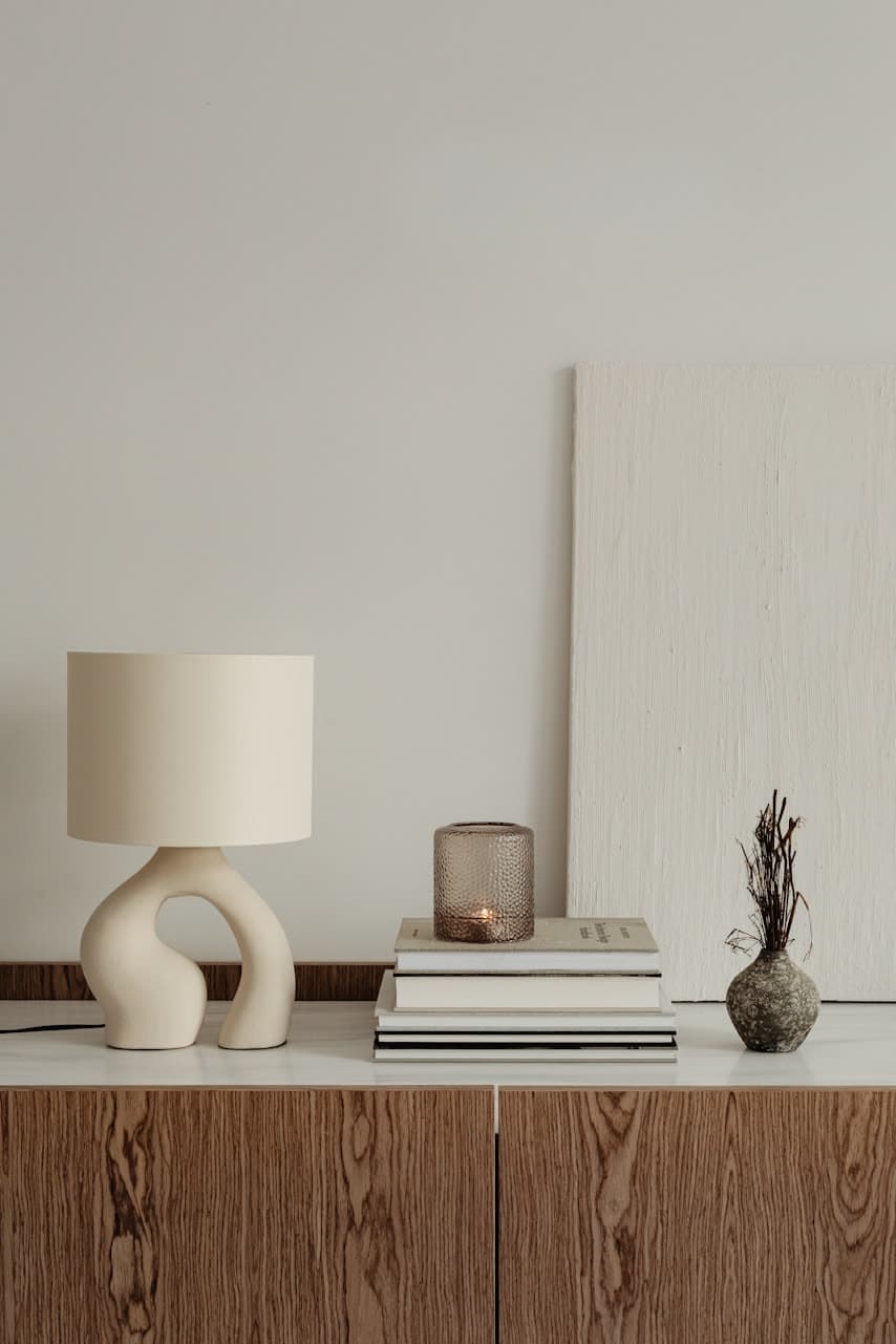 A ceramic table lamp, a mesh candle holder, a stack of books and a dried-flower vase on a pale console, against a plaster wall with a linen canvas leaning on it.
