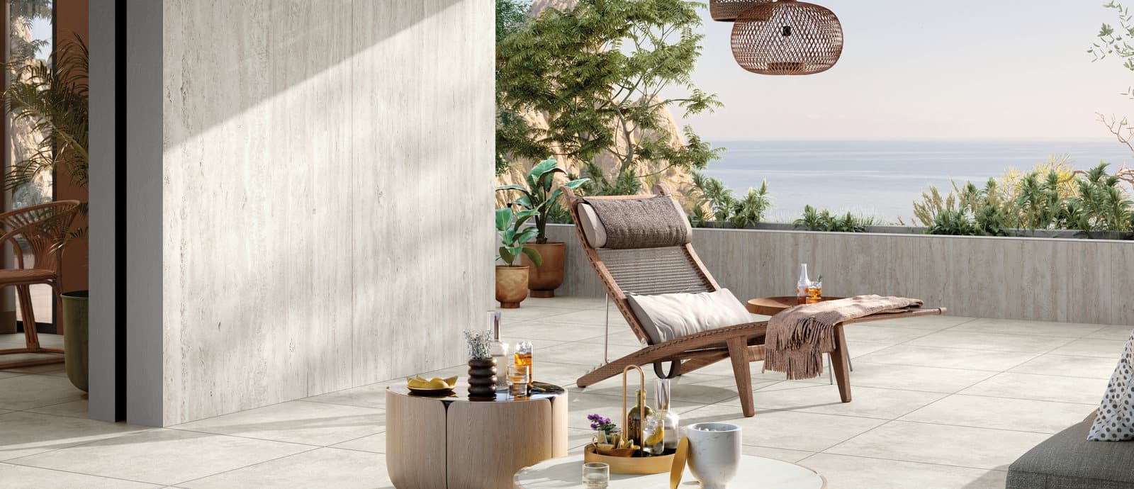 A rattan lounge chair beneath a woven pendant light on a covered stone terrace, looking out over the sea at dusk