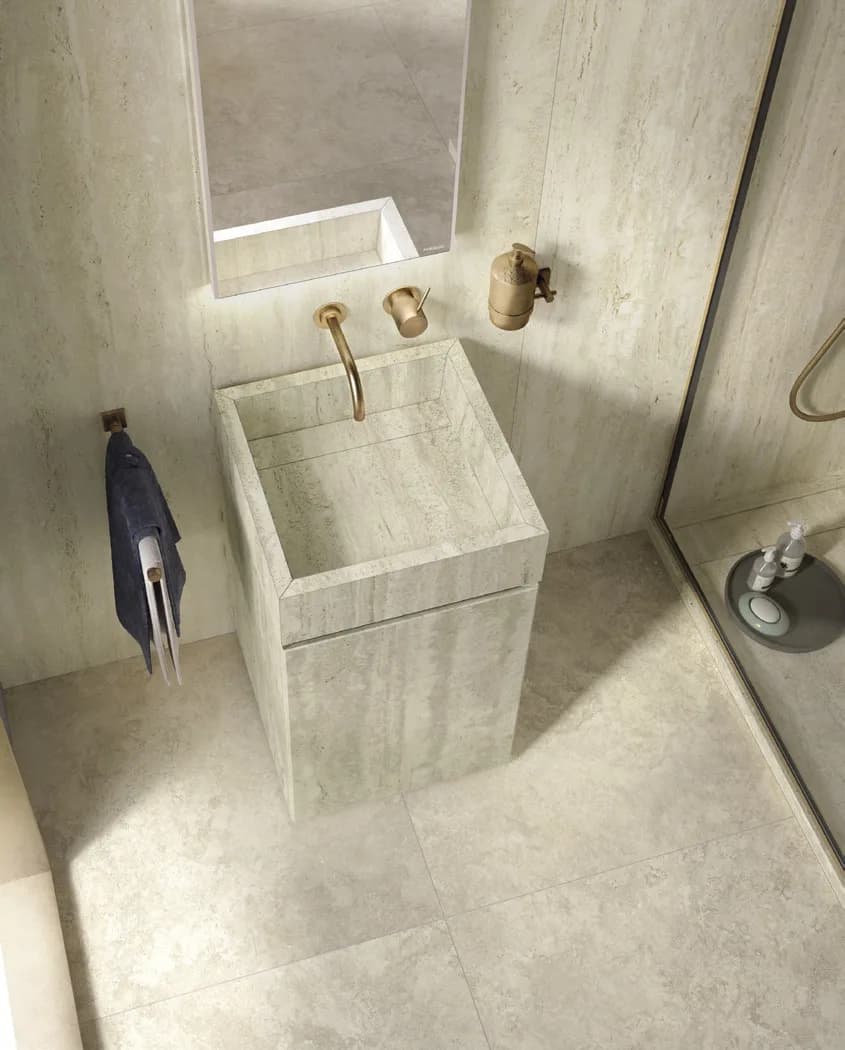 Overhead view of a pale stone pedestal basin in a bathroom corner, with gold fixtures and a dark towel on the wall hook
