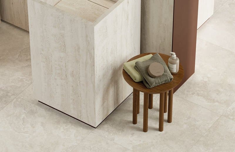 Travertine-clad bathroom corner with a round walnut stool holding folded towels, on large pale stone floor tiles