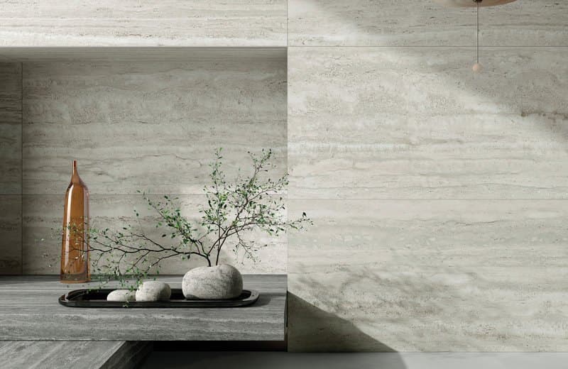 Travertine living-room wall with a recessed alcove holding an amber glass bottle, a small olive tree and pebbles on a dark tray